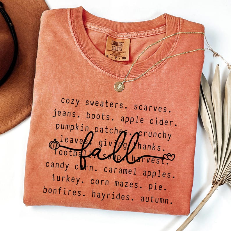 K&ouml;nnte beinhalten: Orangenes Comfort Colors T-Shirt mit einer Liste von W&ouml;rtern zum Thema Herbst in Schwarz, darunter das Wort "fall" in Schreibschrift. Eine goldene Halskette mit Anh&auml;nger liegt auf dem T-Shirt. Das T-Shirt ist auf einer wei&szlig;en Oberfl&auml;che gefaltet.