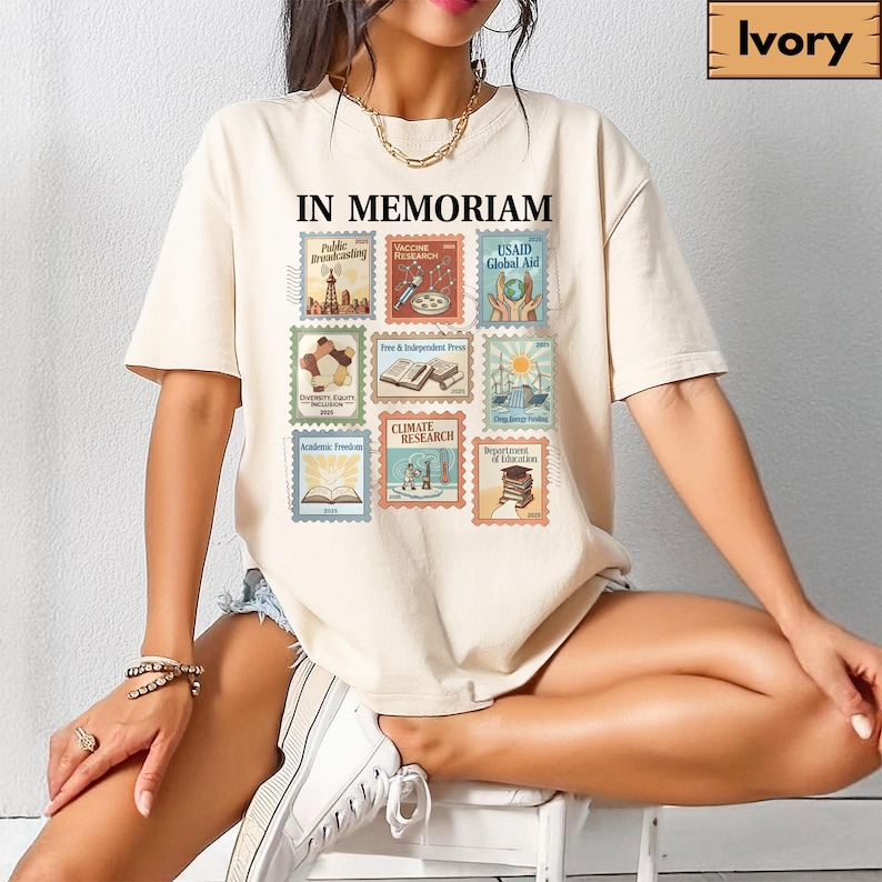 May include: Ivory-colored t-shirt featuring the text "IN MEMORIAM" above a collection of postage stamp-style graphics. The stamps depict various themes, including "Public Broadcasting," "Vaccine Research," and "USAID Global Aid."