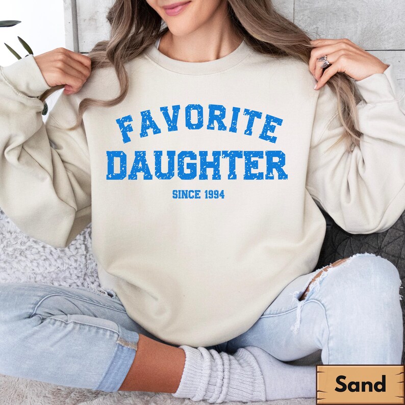 Puede incluir: Una sudadera color arena con el texto azul "FAVORITE DAUGHTER" y "SINCE 1994" debajo. La sudadera es de color neutro y tiene un dise&ntilde;o cl&aacute;sico de cuello redondo. La imagen tambi&eacute;n muestra unos vaqueros azules.