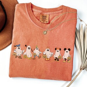 Op de afbeelding: Oranje Comfort Colors T-shirt met een rij cartoongeesten verkleed als Disney-personages, elk met een pompoen. Een gouden ketting met een hanger ligt over het shirt. Het shirt is op een wit oppervlak gevouwen.