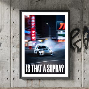 Puede incluir: Póster enmarcado que muestra un coche deportivo plateado derrapando en una calle de la ciudad por la noche. El texto "IS THAT A SUPRA?" está en blanco en la parte inferior. El fondo muestra letreros de neón borrosos.