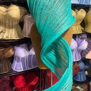 May include: A teal, pleated fabric draped over a mannequin. The fabric is a vibrant turquoise color and appears to be lightweight. The background shows a variety of corsets in different colors, suggesting a fashion or lingerie theme.