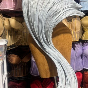 May include: A light blue, pleated fabric draped over a mannequin, creating a flowing, wave-like design. The background features a variety of corsets in shades of pink, brown, red, and yellow, suggesting a lingerie or costume theme.