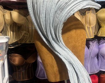 May include: A light blue, pleated fabric draped over a mannequin, creating a flowing, wave-like design. The background features a variety of corsets in shades of pink, brown, red, and yellow, suggesting a lingerie or costume theme.