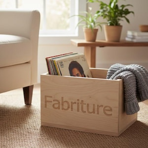 May include: A light-colored wooden storage box with the word "Fabriture" engraved on the front. The box contains vinyl records and a gray knitted blanket. The box is on a woven rug near a white armchair.