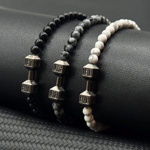 Puede incluir: Tres pulseras de cuentas con colgantes de pesas. Las pulseras presentan cuentas negras, grises y blancas. Los colgantes de pesas son de color plateado y tienen las palabras "LIVE" y "LIFT" grabadas. Las pulseras se exhiben sobre una superficie negra.
