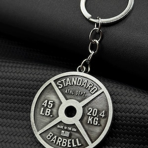 Op de afbeelding: Zilverkleurige metalen sleutelhanger in de vorm van een gewichtsplaat. Er staat "STANDARD ATHL DEPT. 45 LB. 20.4 KG. MADE IN THE USA BARBELL" op.