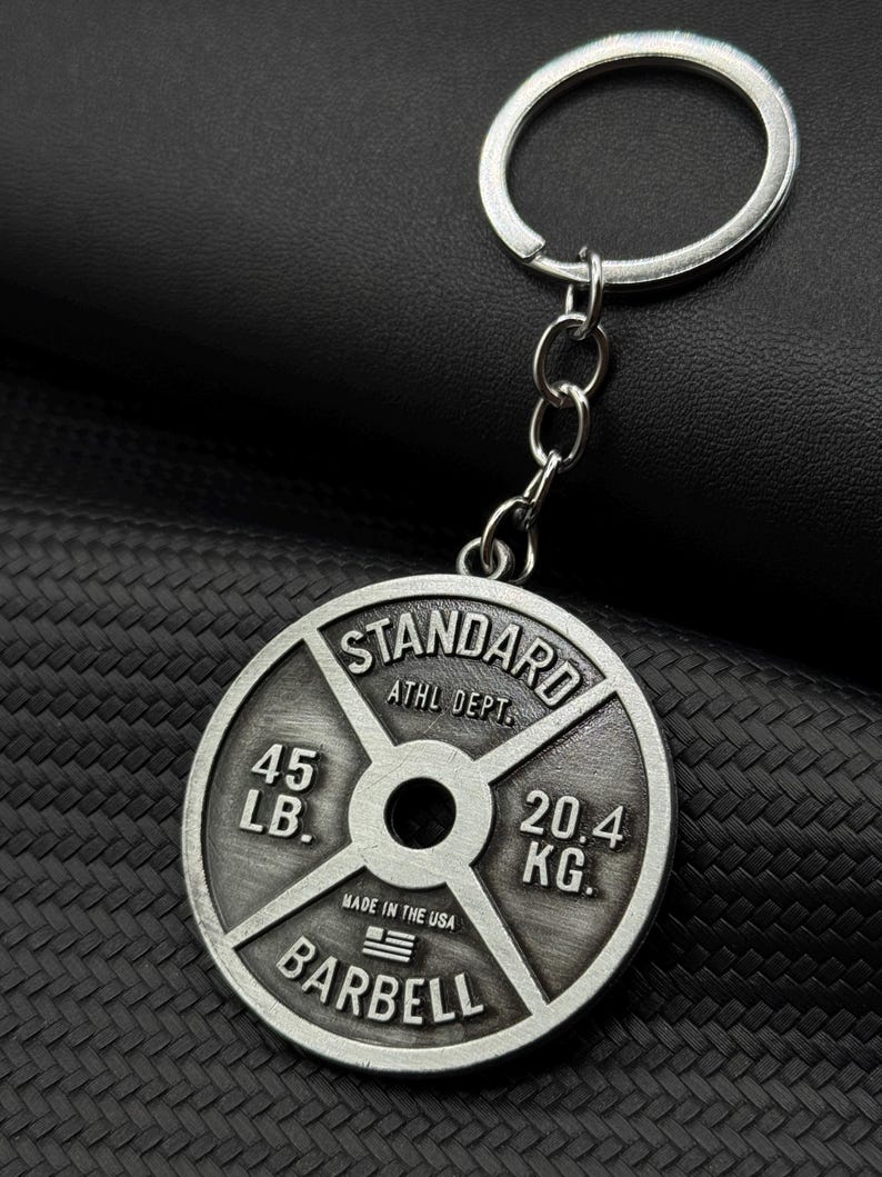 Op de afbeelding: Zilverkleurige metalen sleutelhanger in de vorm van een gewichtsplaat. Er staat "STANDARD ATHL DEPT. 45 LB. 20.4 KG. MADE IN THE USA BARBELL" op.