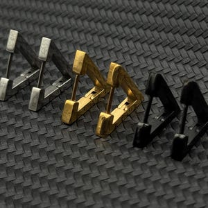 May include: Five pairs of triangle-shaped earrings in silver, gold, and black metal. The earrings are arranged in a row on a black textured surface.