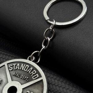 Op de afbeelding: Zilveren sleutelhanger met een gewichtsplaatbedel. De plaat is grijs en heeft de tekst "STANDARD ATHL DEPT. 20.4 KG" erop gedrukt.
