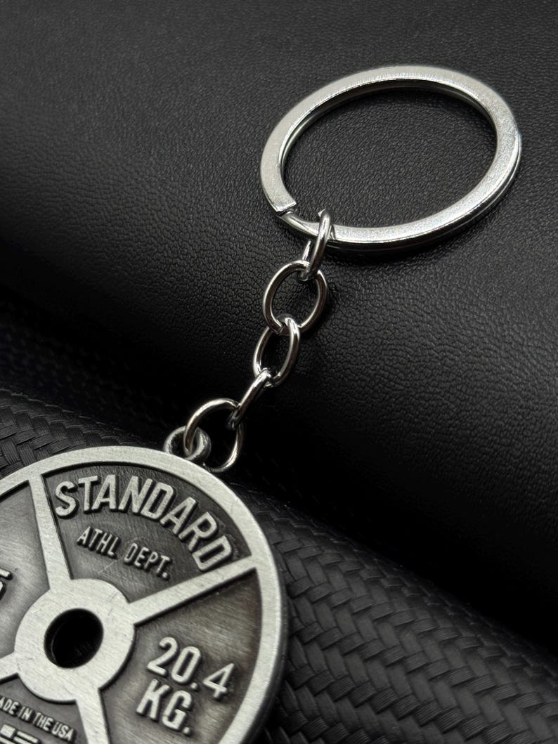 Op de afbeelding: Zilveren sleutelhanger met een gewichtsplaatbedel. De plaat is grijs en heeft de tekst "STANDARD ATHL DEPT. 20.4 KG" erop gedrukt.