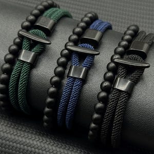 May include: Three beaded bracelets with black beads and braided rope in green, blue, and black. Each bracelet features a metal clasp and a decorative metal accent. The bracelets are displayed on a dark, textured surface.