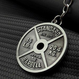 Op de afbeelding: Zilverkleurige metalen sleutelhanger in de vorm van een gewichtsplaat. Er staat "STANDARD ATHL DEPT. 45 LB. 20.4 KG. BARBELL" en "MADE IN THE USA" met een kleine Amerikaanse vlag.