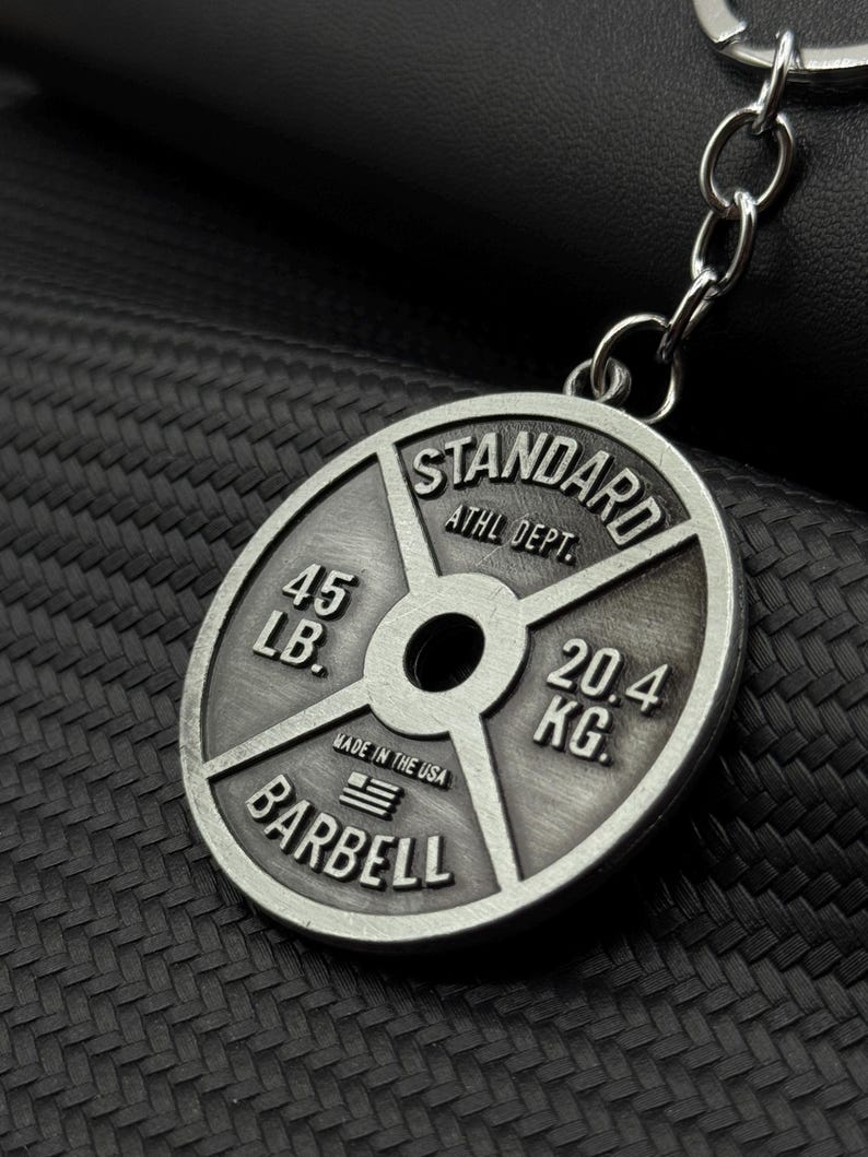 Op de afbeelding: Zilverkleurige metalen sleutelhanger in de vorm van een gewichtsplaat. Er staat "STANDARD ATHL DEPT. 45 LB. 20.4 KG. BARBELL" en "MADE IN THE USA" met een kleine Amerikaanse vlag.