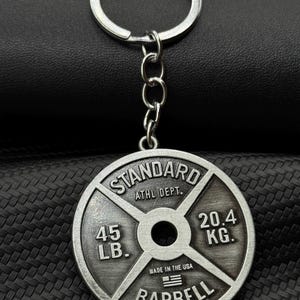 Op de afbeelding: Zilverkleurige metalen sleutelhanger in de vorm van een gewichtsplaat. De plaat heeft de tekst "STANDARD" bovenaan, "ATHL DEPT." in het midden, "45 LB." links, "20.4 KG." rechts, "MADE IN THE USA" met een kleine Amerikaanse vlag eronder en "BARBELL" onderaan.