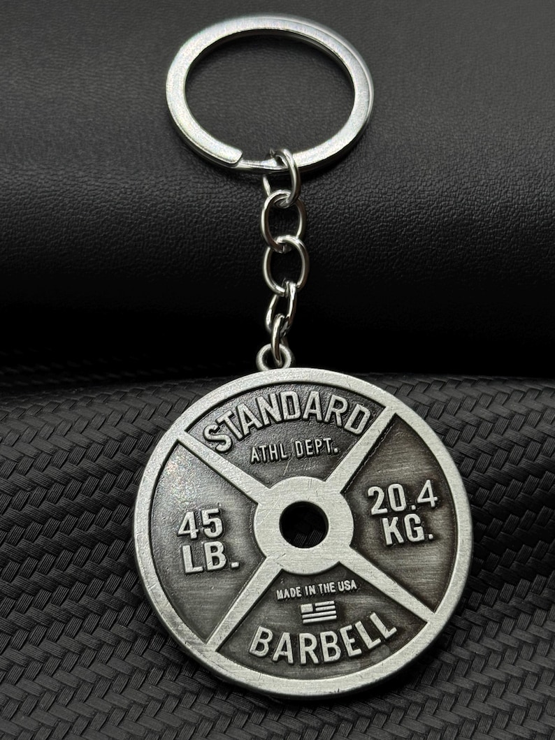 Op de afbeelding: Zilverkleurige metalen sleutelhanger in de vorm van een gewichtsplaat. De plaat heeft de tekst "STANDARD" bovenaan, "ATHL DEPT." in het midden, "45 LB." links, "20.4 KG." rechts, "MADE IN THE USA" met een kleine Amerikaanse vlag eronder en "BARBELL" onderaan.