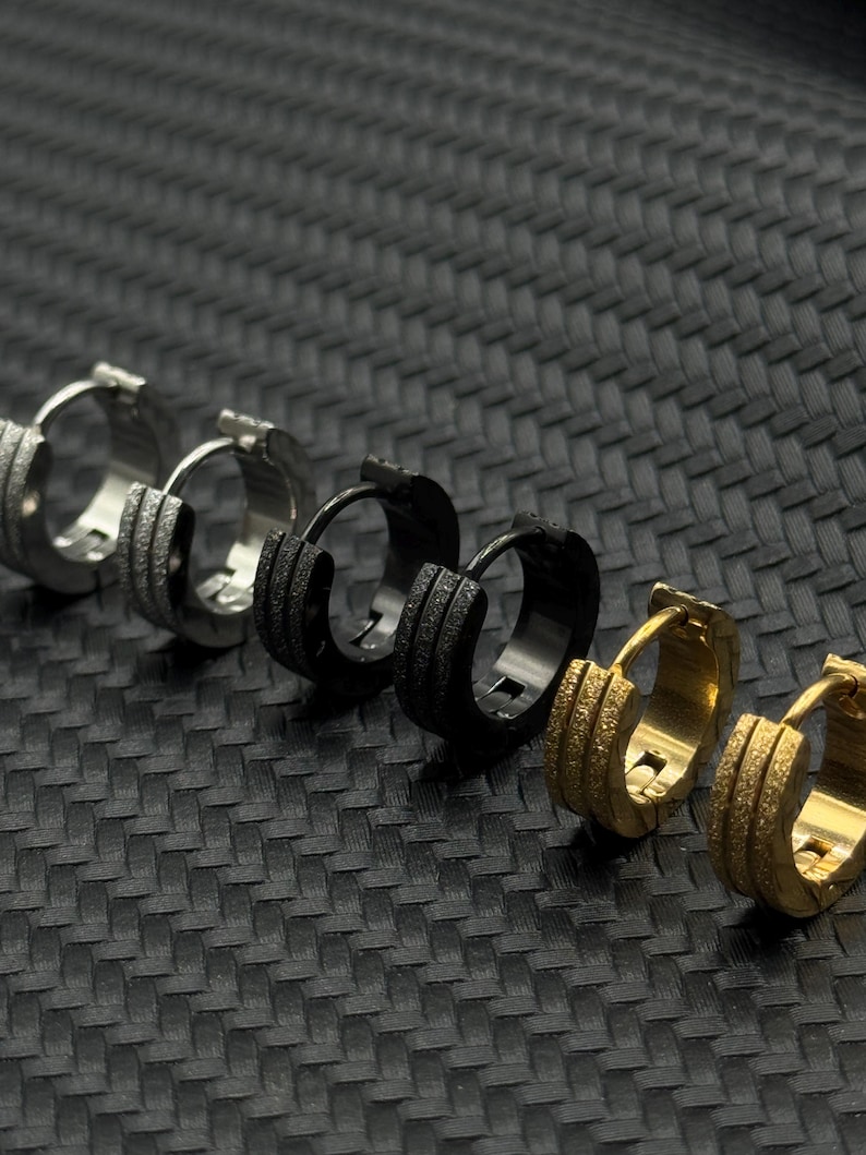 May include: A set of six hoop earrings in different finishes, including silver, black, and gold. The earrings have a textured surface and are arranged in a row.