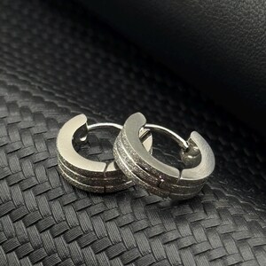 May include: A pair of silver hoop earrings with a textured finish. The earrings are on a black textured surface.