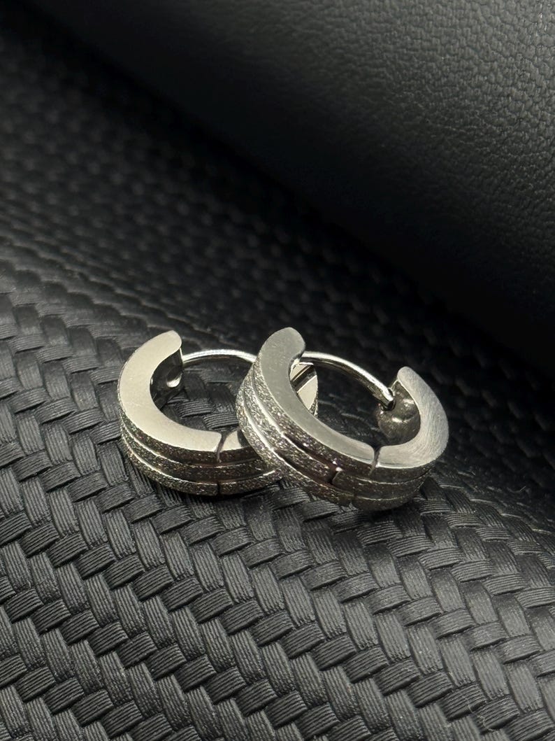 May include: A pair of silver hoop earrings with a textured finish. The earrings are on a black textured surface.