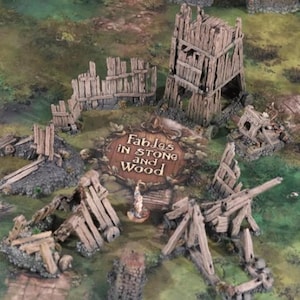 May include: A tabletop game scene featuring miniature wooden structures and siege weapons. The scene includes a central sign that reads "Fables in Stone and Wood". The structures are weathered and detailed, set against a green and brown terrain.