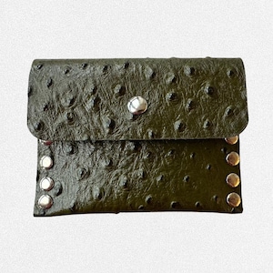 Puede incluir: Un estuche rectangular de color verde oliva con una superficie texturizada y rugosa que recuerda a la piel de avestruz. La solapa cuenta con un cierre de presión metálico plateado y los laterales están adornados con tres tachuelas plateadas.