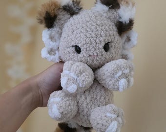 May include: A plush toy crafted from beige, brown, and white fuzzy yarn. The creature features large black eyes, prominent ears with multi-colored patches, and white accents on the paws and head. It has a long, thick, multi-colored tail.