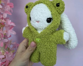 May include: A handmade plush bunny wearing a green frog costume. The bunny has white fur, large black eyes, and a pink nose. The frog costume features a hood with frog eyes and a green button closure. The background is a soft purple.