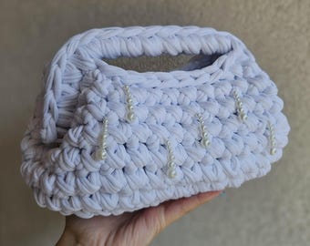 May include: A white, hand-crocheted clutch bag with a braided handle. The bag is adorned with dangling pearl accents. The texture of the crochet is visible, and the bag is held in a hand.