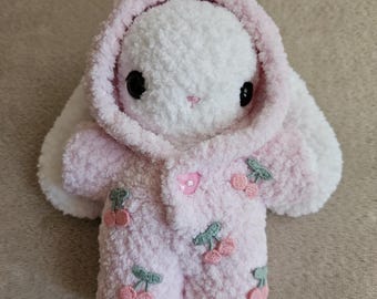 May include: A plush white bunny wearing a pink hooded jumpsuit adorned with embroidered cherries. The bunny has large black eyes and a pink nose. The jumpsuit has a single pink button closure. The bunny's ears are white.