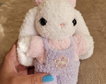 May include: A handmade white plush bunny with a pink bow, black eyes, and a pink nose. The bunny wears a pink and lavender outfit with a flower applique. The plush toy is soft and cuddly.