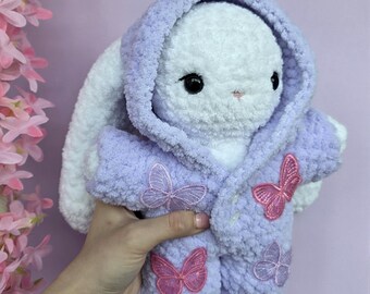 May include: A white plush bunny wearing a lavender hooded jacket with pink butterfly appliqués. The bunny has black eyes and a pink nose. The jacket is a soft, textured material. The bunny is a cute, handmade item.