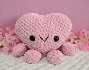 May include: A handmade pink crocheted heart-shaped plush toy with black safety eyes and a stitched smile. The plush has four small, rounded appendages and is displayed on a white, textured surface with pink flowers in the background.