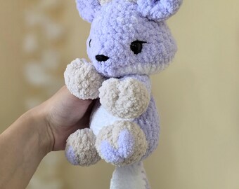 May include: A plush stuffed animal made of soft lavender and beige yarn. The creature features large ears, small horns, a white belly, and a long tail. It has black embroidered eyes and a small black nose, with a textured, fuzzy appearance throughout.