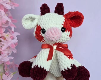May include: A handmade crochet cow plushie with a red and white pattern. The plushie has a pink nose, black eyes, and a red bow. The cow is approximately 8 inches tall.
