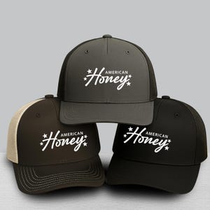 May include: Three trucker hats in shades of gray, brown, and black. Each hat features the text "American Honey" in white script with star accents. The hats have mesh backs and curved brims.