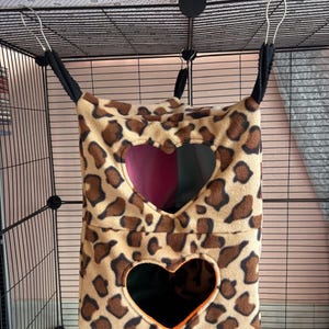 Puede incluir: Una hamaca colgante para mascotas de dos niveles hecha de tela polar con estampado de leopardo en tonos beige y marrón. La estructura incluye dos aberturas en forma de corazón en el frente, correas de tela negra y ganchos metálicos.