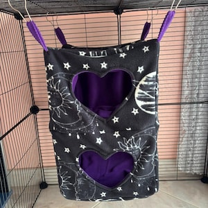Peut inclure: Un hamac suspendu pour animal de compagnie à deux niveaux en tissu polaire noir avec un motif céleste blanc représentant des soleils, des lunes et des étoiles. Il possède deux ouvertures en forme de cœur et un intérieur violet uni.