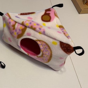 Peut inclure: Une cachette pour animal en polaire triangulaire avec une base blanche et un motif répétitif de beignets roses et marron avec des pépites jaunes. La structure présente une ouverture circulaire avec un intérieur rose et des boucles noires.