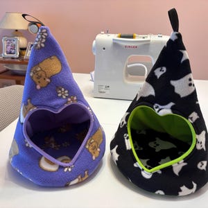 Peut inclure: Deux abris pour animaux en polaire en forme de cône avec des ouvertures en forme de cœur. L'un est violet avec un motif de cochons d'Inde marron, l'autre est noir avec des fantômes blancs et une bordure intérieure vert citron.