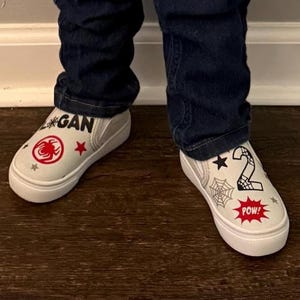 May include: White slip-on sneakers with a red spider emblem, stars, and the name "Logan" printed on one shoe. The other shoe features a number "2", a spider web, and the word "POW!" in a red burst.