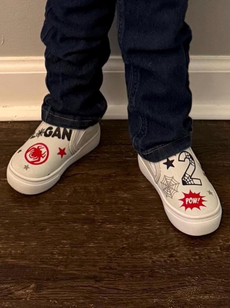 May include: White slip-on sneakers with a red spider emblem, stars, and the name "Logan" printed on one shoe. The other shoe features a number "2", a spider web, and the word "POW!" in a red burst.