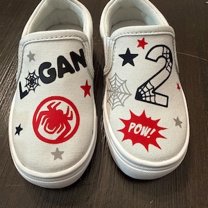 May include: A pair of light gray slip-on shoes with white soles. One shoe features the name "LOGAN" and a red spider emblem. The other shoe has the number "2" and a "POW!" speech bubble.