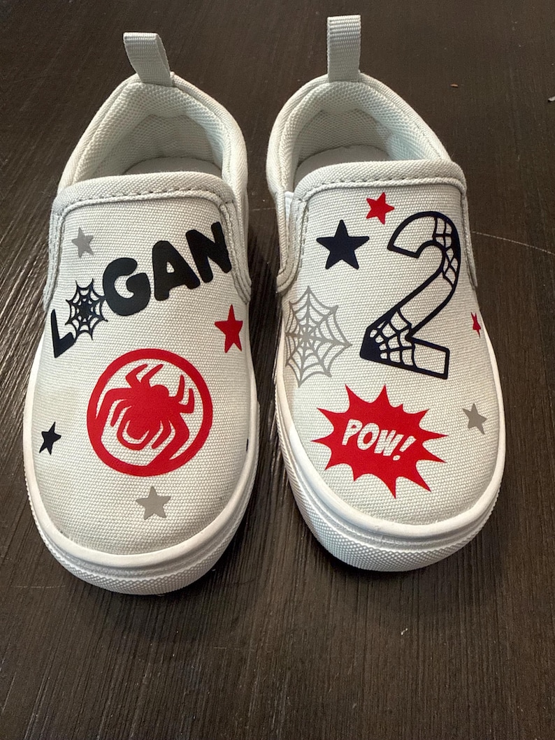 May include: A pair of light gray slip-on shoes with white soles. One shoe features the name "LOGAN" and a red spider emblem. The other shoe has the number "2" and a "POW!" speech bubble.