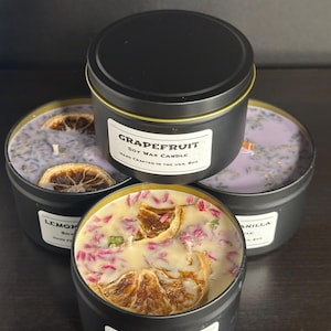 May include: Four black metal tins containing soy wax candles. The front candle is topped with dried orange slices and pink petals. The other candles feature lavender-colored wax with dried citrus slices and herbs. Each tin has a white label.