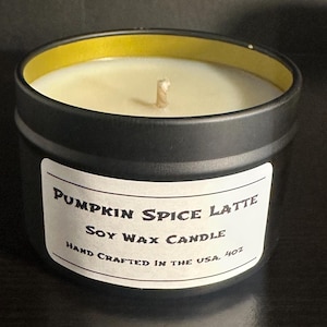 May include: A black metal tin candle with a white label featuring the text Pumpkin Spice Latte Soy Wax Candle Hand Crafted in the USA 4oz. The candle has a single centered wick and off-white wax inside a container with a gold-colored inner rim.