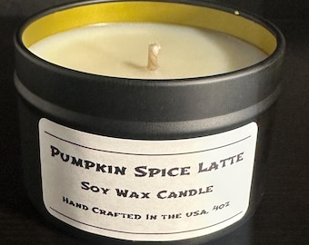 May include: A black metal tin candle with a white label featuring the text Pumpkin Spice Latte Soy Wax Candle Hand Crafted in the USA 4oz. The candle has a single centered wick and off-white wax inside a container with a gold-colored inner rim.