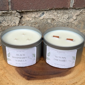 May include: Two round metal tins containing soy wax candles. The left candle has three wicks and a label reading The Thrifty Wick, Black Raspberry & Vanilla, Soy Wax Candle. The right candle has two wooden wicks and a label reading The Thrifty Wick, Autumn Orchard, Soy Wax Candle.