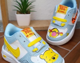 Könnte beinhalten: Ein Paar hellblaue und weiße Air Force 1 Sneaker mit Winnie-Puuh-Grafiken. Die Schuhe haben gelbe Akzente, darunter der Nike-Swoosh und die Schnürsenkel. Der Text "Winnie the Pooh Air Force 1" ist auf der Zunge aufgedruckt.