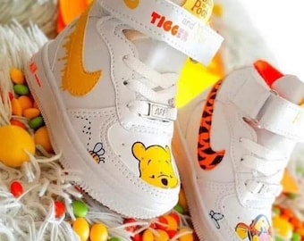 Könnte beinhalten: Weiße High-Top-Sneaker mit gelbem Swoosh und Winnie Puuh und Tigger-Designs. Die Schuhe sind mit Comicfiguren und Bienen verziert.