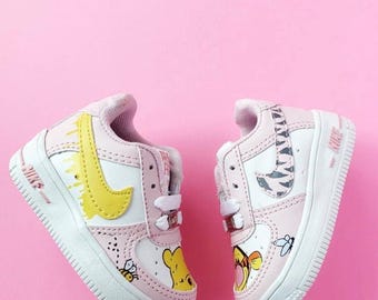 Könnte beinhalten: Paar rosa-weiße Nike Air Force 1 Babyschuhe. Die Schuhe haben gelbe Swooshes, Cartoon-Charakter-Grafiken und eine weiße Sohle. Das Design umfasst eine Biene und Winnie Puuh.
